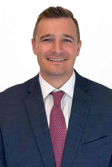 Man wearing a blue suit, white shirt, and red patterned tie, smiling at the camera against a plain white background.