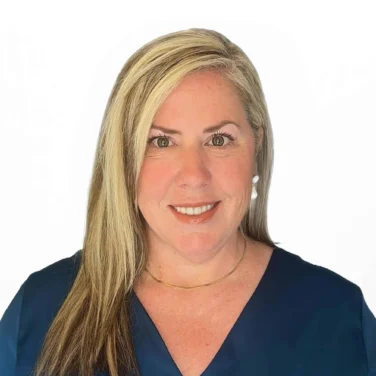 A woman financial advisor with blonde hair, wearing a navy blue blouse, gold necklace, and pearl earrings, smiles at the camera against a plain white background.