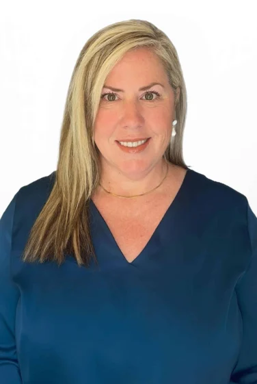 A woman financial advisor with blonde hair, wearing a navy blue blouse, gold necklace, and pearl earrings, smiles at the camera against a plain white background.