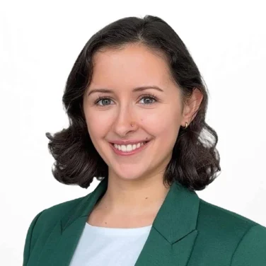Woman with short dark hair wearing a green blazer and white shirt, smiling, standing against a plain white background.