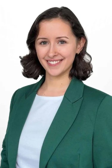 Woman with short dark hair wearing a green blazer and white shirt, smiling, standing against a plain white background.