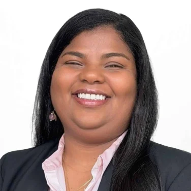 Woman with long straight black hair wearing a black blazer, light pink shirt, and necklace, smiling at the camera against a plain white background.