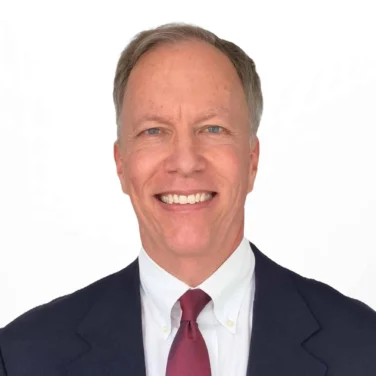 A man in a dark suit, white shirt, and red tie is smiling at the camera against a plain white background.