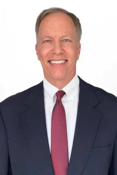 A man in a dark suit, white shirt, and red tie is smiling at the camera against a plain white background.