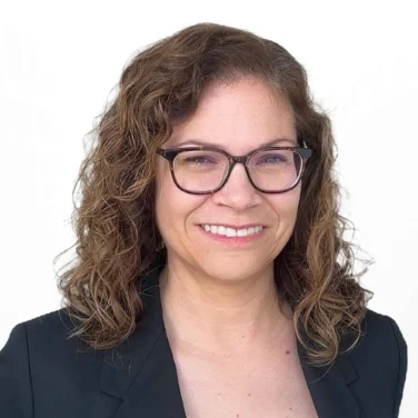 Woman with curly brown hair and glasses wearing a black blazer and light top, smiling against a plain white background.