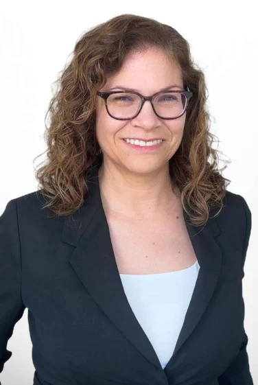 Woman with curly brown hair and glasses wearing a black blazer and light top, smiling against a plain white background.