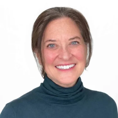 Woman with brown hair and blue eyes wearing a teal turtleneck sweater, smiling at the camera against a white background.
