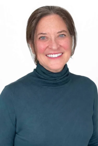 Woman with brown hair and blue eyes wearing a teal turtleneck sweater, smiling at the camera against a white background.