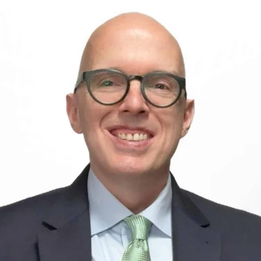 Bald man with glasses wearing a dark suit, light blue shirt, and light green tie, smiling at the camera against a plain white background.