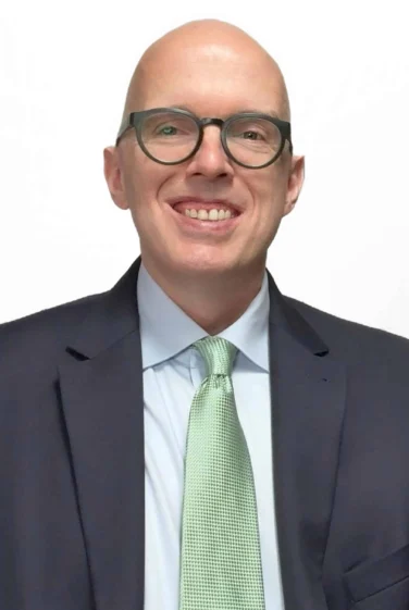 Bald man with glasses wearing a dark suit, light blue shirt, and light green tie, smiling at the camera against a plain white background.
