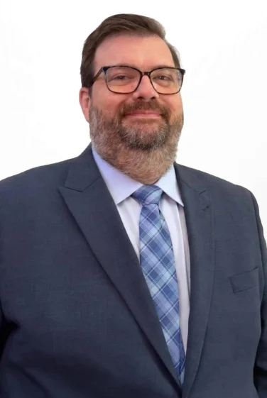 A man in a dark suit, blue patterned tie, and glasses stands against a plain white background, smiling slightly.