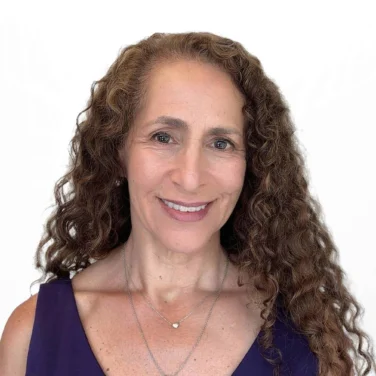 Woman with long curly hair wearing a sleeveless purple top and two necklaces, smiling against a plain white background.