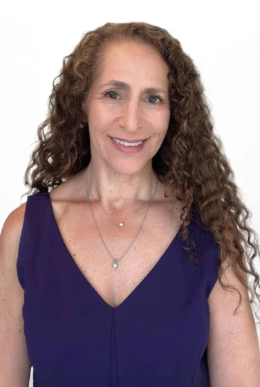 Woman with long curly hair wearing a sleeveless purple top and two necklaces, smiling against a plain white background.