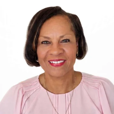 A woman with short dark hair, wearing a light pink blouse and a necklace, smiles at the camera against a plain white background.