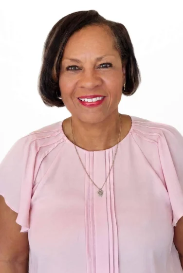 A woman with short dark hair, wearing a light pink blouse and a necklace, smiles at the camera against a plain white background.