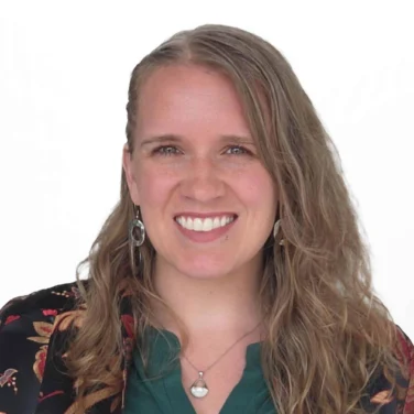 A woman with long, wavy light brown hair is smiling, wearing a green top with gold buttons and a floral-patterned blazer. The background is plain white.