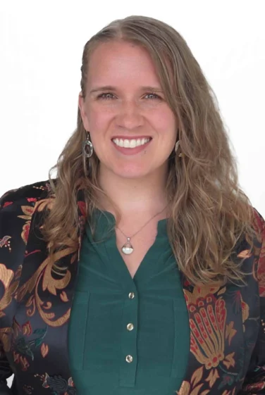 A woman with long, wavy light brown hair is smiling, wearing a green top with gold buttons and a floral-patterned blazer. The background is plain white.