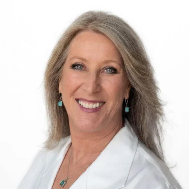 Smiling adult woman with long gray hair, wearing a white collared shirt and turquoise jewelry, posed against a plain white background.
