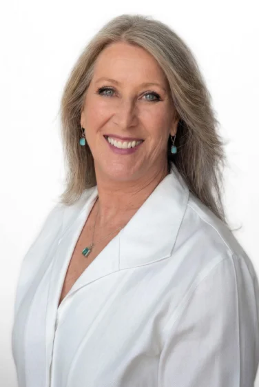 Smiling adult woman with long gray hair, wearing a white collared shirt and turquoise jewelry, posed against a plain white background.