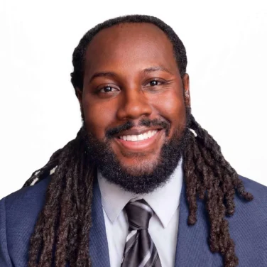 A man in a blue suit, light shirt, and striped tie smiles at the camera against a plain white background. He has a beard and long, dark dreadlocks.