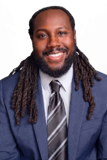 A man in a blue suit, light shirt, and striped tie smiles at the camera against a plain white background. He has a beard and long, dark dreadlocks.