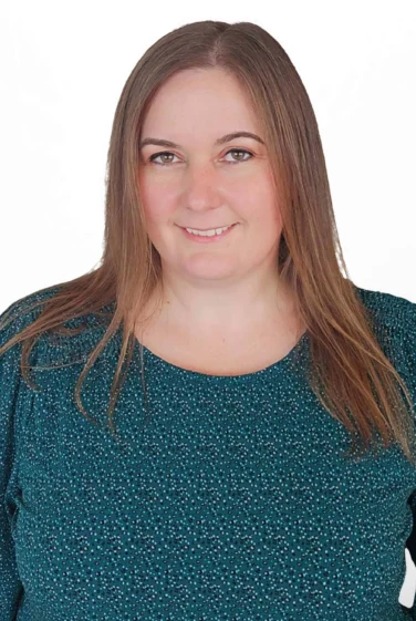 Woman with straight light brown hair wearing a green patterned top, smiling slightly, standing against a plain white background.
