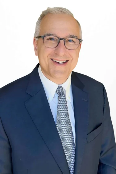 A man wearing glasses, a navy suit, white shirt, and patterned tie smiles at the camera against a plain white background.