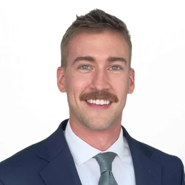 Man in a navy suit, white shirt, and light blue tie, with light brown hair and a mustache, smiling at the camera against a plain white background.