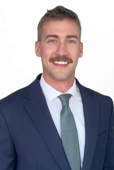 Man in a navy suit, white shirt, and light blue tie, with light brown hair and a mustache, smiling at the camera against a plain white background.
