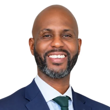 A man with a bald head, dark beard, and mustache smiles while wearing a blue suit, white shirt, and green tie, standing against a white background.