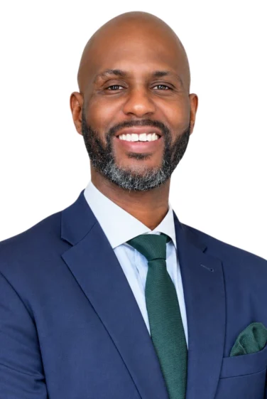 A man with a bald head, dark beard, and mustache smiles while wearing a blue suit, white shirt, and green tie, standing against a white background.
