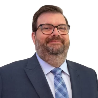 A man in a blue suit, light blue shirt, and plaid tie stands against a white background, facing forward and smiling slightly.