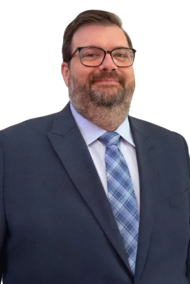 A man in a blue suit, light blue shirt, and plaid tie stands against a white background, facing forward and smiling slightly.