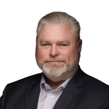 A man with gray hair and a beard in a dark suit jacket and light-colored shirt poses against a plain white background.