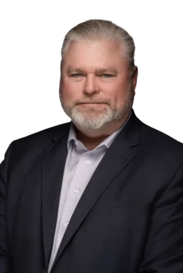 A man with gray hair and a beard in a dark suit jacket and light-colored shirt poses against a plain white background.