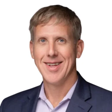 Man with short light brown hair wearing a navy blue suit jacket and light purple dress shirt, smiling, against a plain white background.