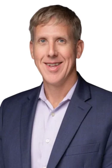 Man with short light brown hair wearing a navy blue suit jacket and light purple dress shirt, smiling, against a plain white background.