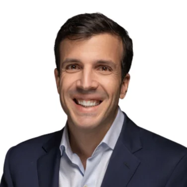 Man in a navy suit jacket and light blue shirt smiling at the camera against a white background.