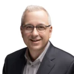 A man with short gray hair and glasses is wearing a dark suit jacket over a light-colored shirt, smiling at the camera against a white background.
