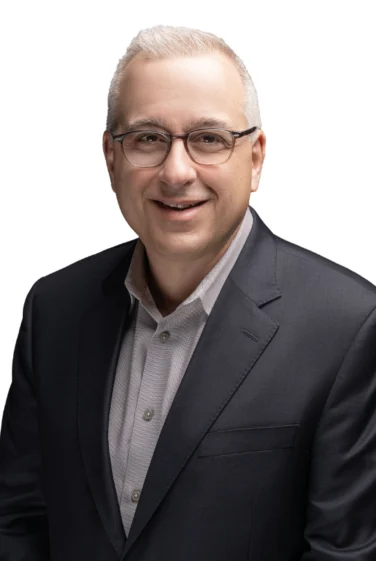 A man with short gray hair and glasses is wearing a dark suit jacket over a light-colored shirt, smiling at the camera against a white background.