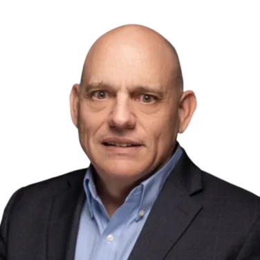 A bald man wearing a dark suit jacket and light blue collared shirt poses against a plain white background.