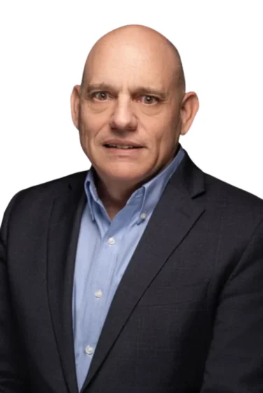 A bald man wearing a dark suit jacket and light blue collared shirt poses against a plain white background.