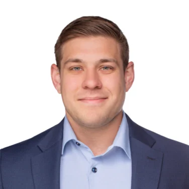 Man in a blue suit jacket and light blue dress shirt, standing against a plain white background, looking at the camera with a neutral expression.