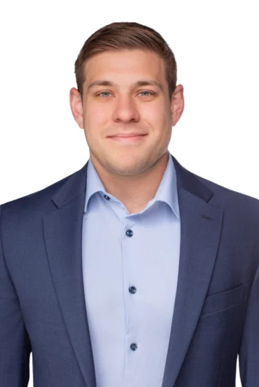 Man in a blue suit jacket and light blue dress shirt, standing against a plain white background, looking at the camera with a neutral expression.