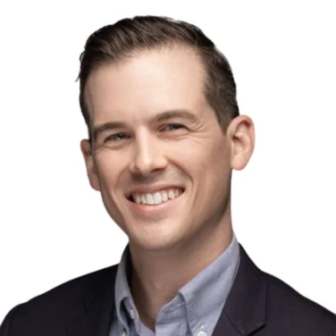 Man with short brown hair in a dark blazer and light button-up shirt smiling against a plain white background.