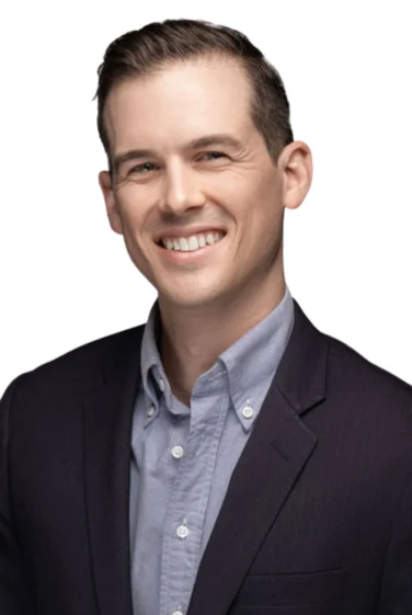 Man with short brown hair in a dark blazer and light button-up shirt smiling against a plain white background.