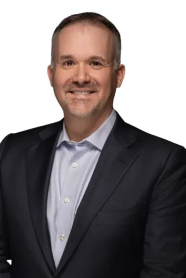 Man wearing a dark suit jacket and light blue dress shirt, smiling at the camera, with a plain white background.