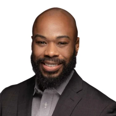 Smiling man with a beard wearing a dark suit jacket and a light-colored shirt, posed against a white background.