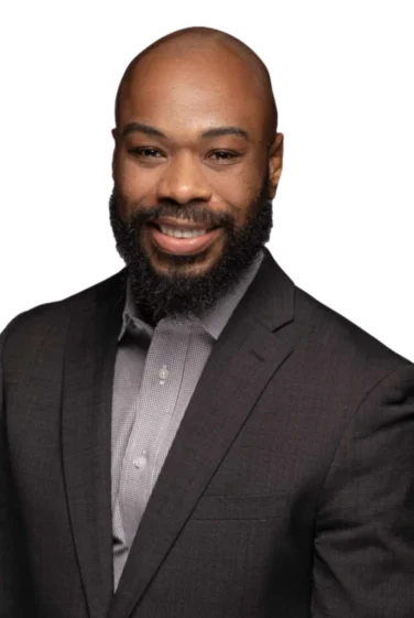 Smiling man with a beard wearing a dark suit jacket and a light-colored shirt, posed against a white background.