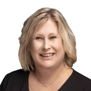 Smiling middle-aged woman with blonde hair wears a black top and a small necklace, posing against a plain white background.
